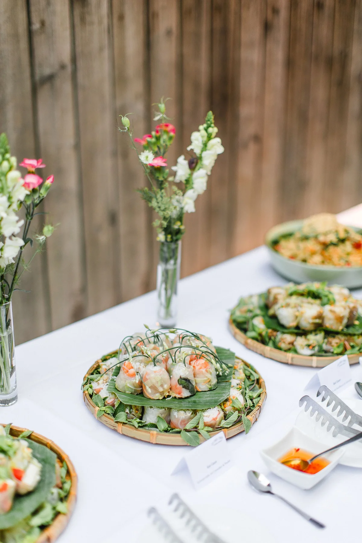 Vietnamesische Sommerrollen auf Bambusteller mit frischen Blumen auf weisser Tischwäsche beim Outdoor-Catering